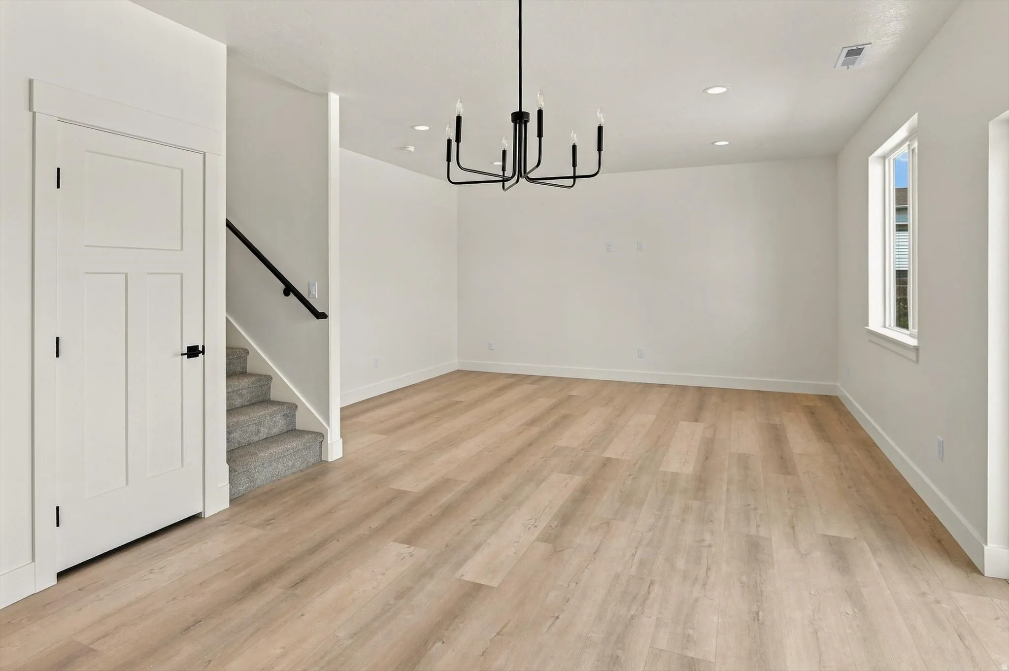 Recessed lighting, wood-finish flooring, white walls, black chandelier, and a window with white trim