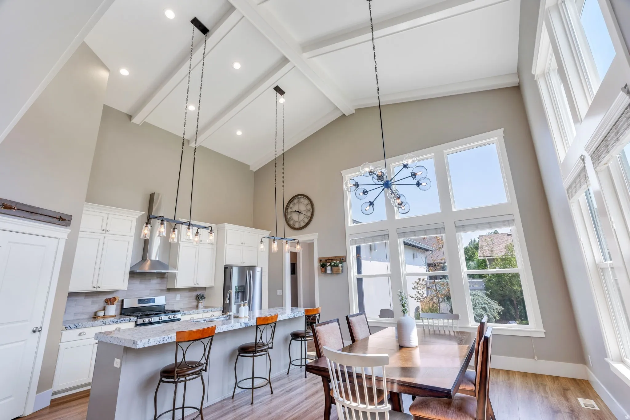 Soaring vaulted ceiling with exposed beams, expansive window wall, recessed lighting, wood-finish flooring, and a kitchen island with stone countertops