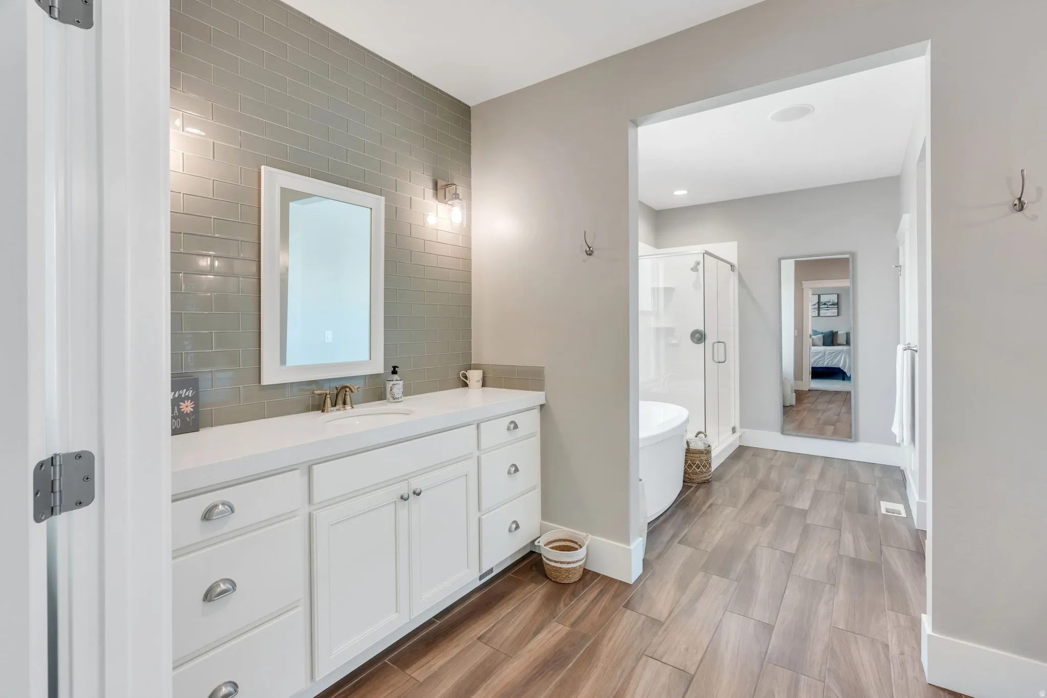 Dual vanity with white cabinetry and chrome hardware, subway tile accent wall, wood-finish flooring, glass-enclosed shower, and freestanding soaking tub
