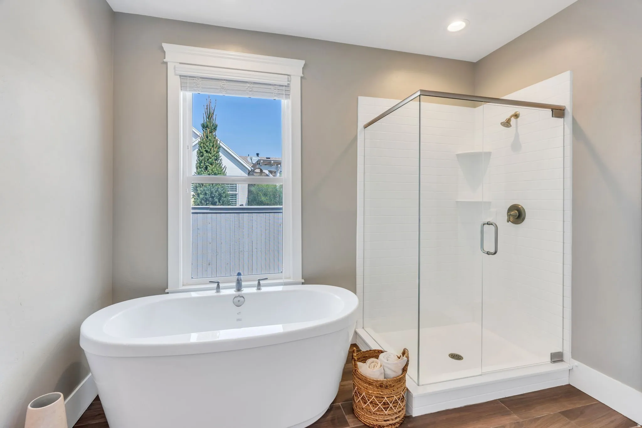 Freestanding soaking tub with polished chrome fixtures, glass-enclosed shower with subway tile surround, wood-finish flooring, and recessed lighting