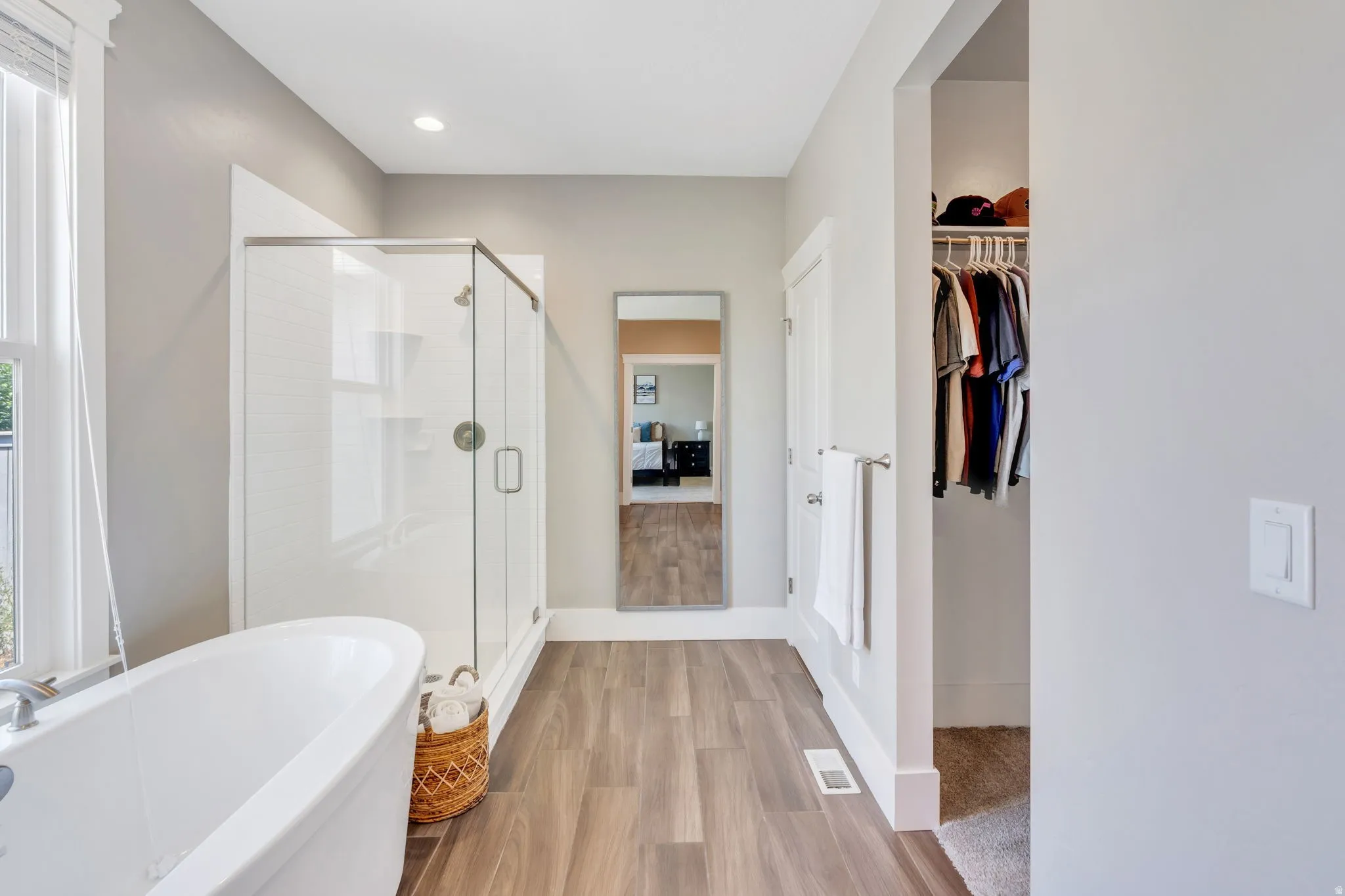 Freestanding soaking tub, glass-enclosed shower, wood-finish flooring, recessed lighting, and an open closet with built-in shelving