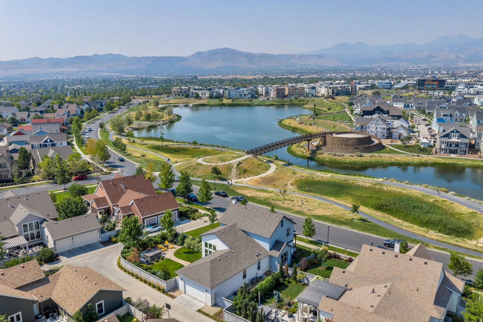 Expansive urban landscape featuring a large lake, surrounding greenery, pedestrian bridge, and distant mountains