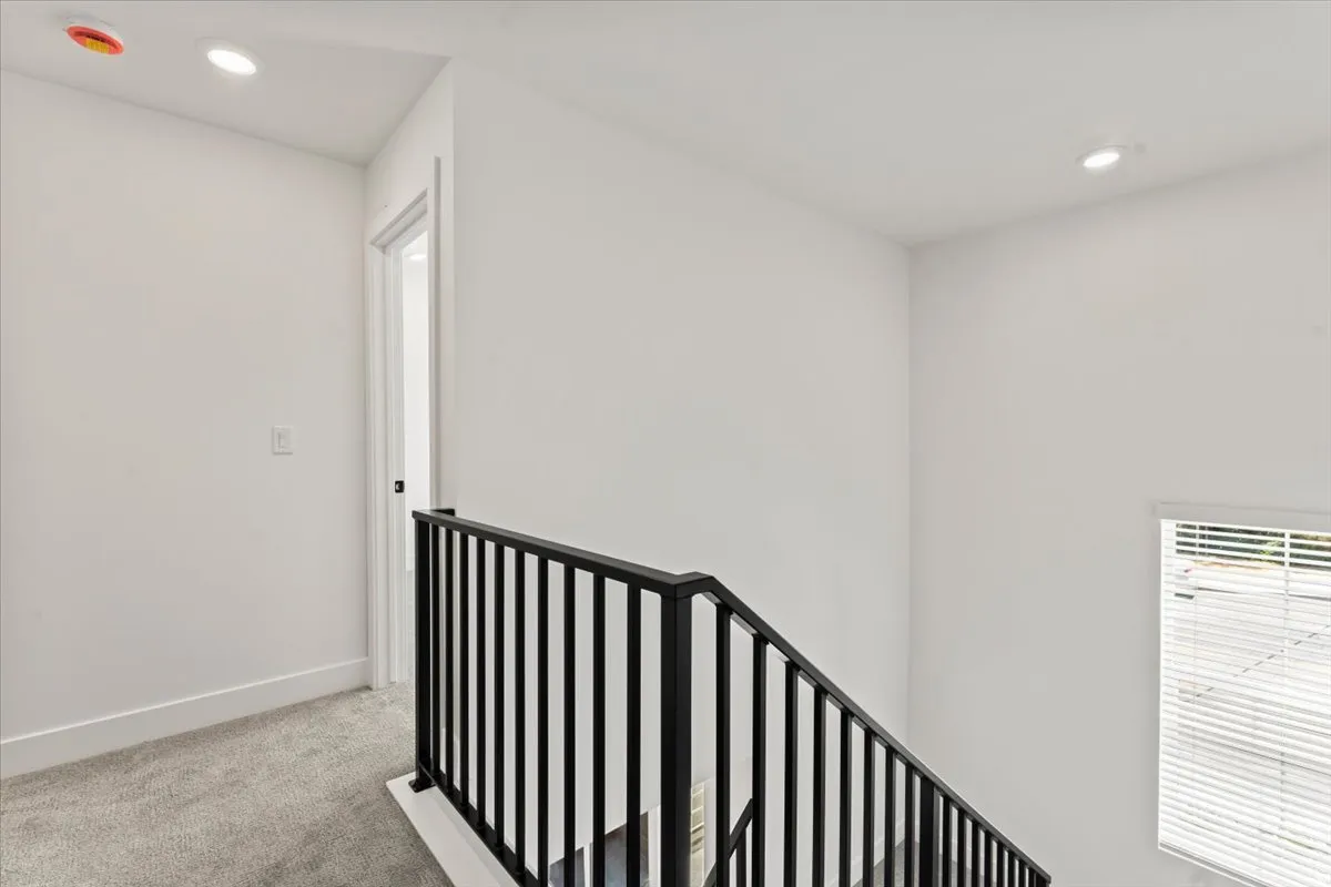 White walls, recessed lighting, carpeted hallway, black metal stair railing, and a window with horizontal blinds