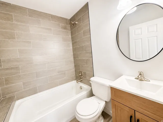 Bathtub with horizontal tile surround, wood-finish vanity with white countertop, round wall-mounted mirror, and brushed nickel fixtures