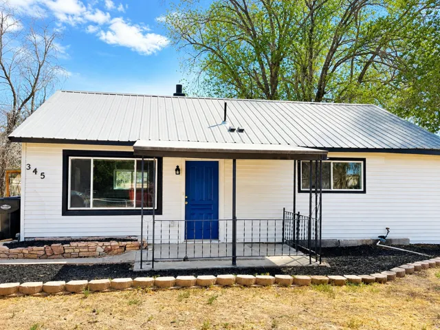 White siding exterior with a metal roof, blue entry door, black-framed windows, and a covered porch with wrought iron railing