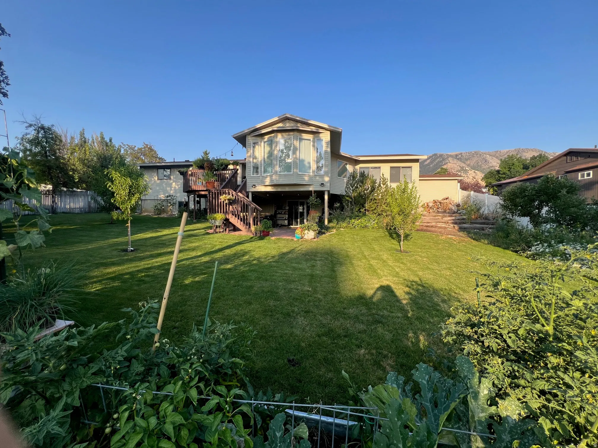 Expansive backyard featuring a manicured lawn, mature landscaping with established trees and shrubs, and wooden deck with stairs leading to the elevated residence