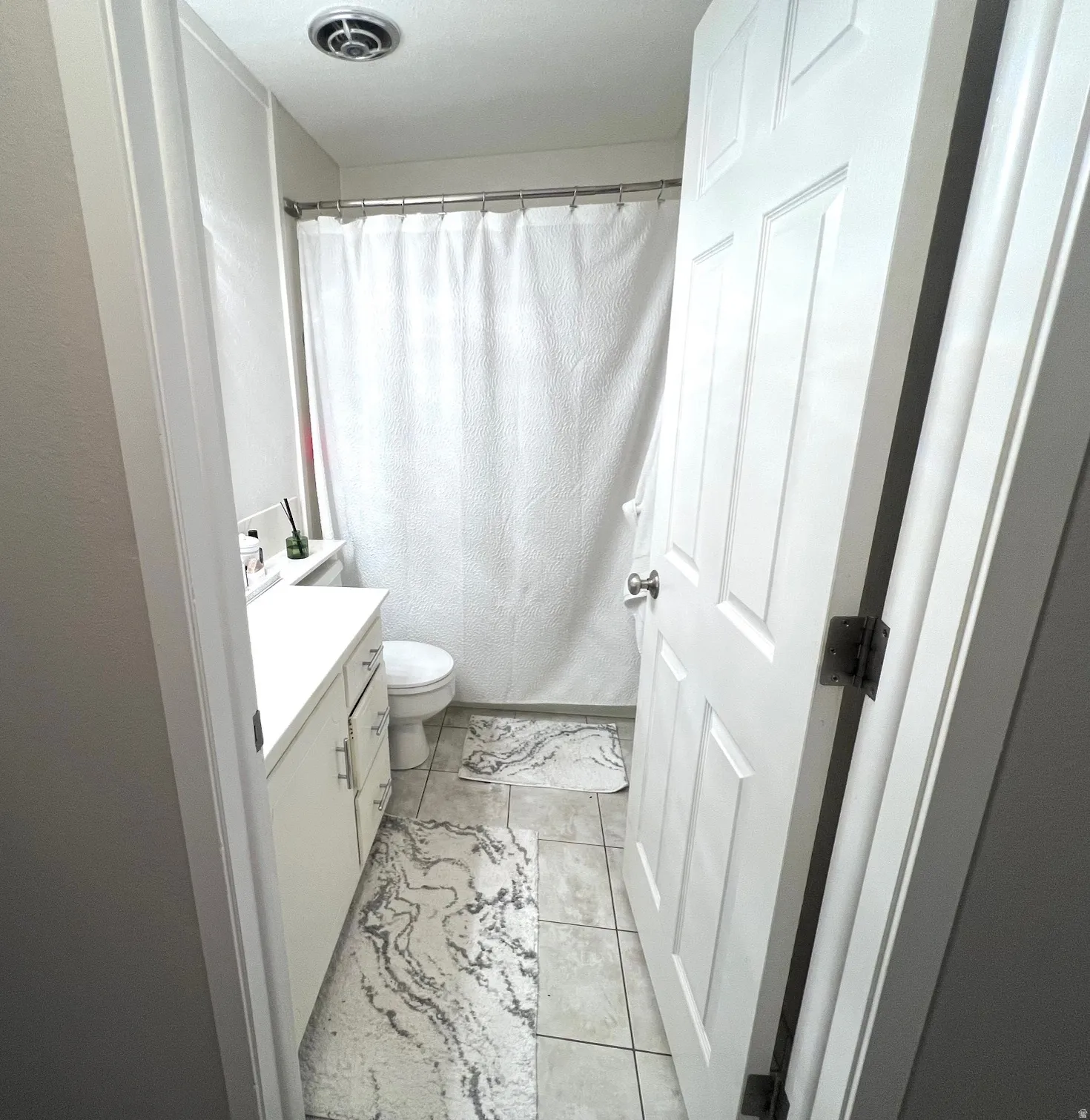 White paneled door, light-colored tile flooring, white vanity with cabinet storage, white shower curtain, and a ceiling-mounted exhaust fan