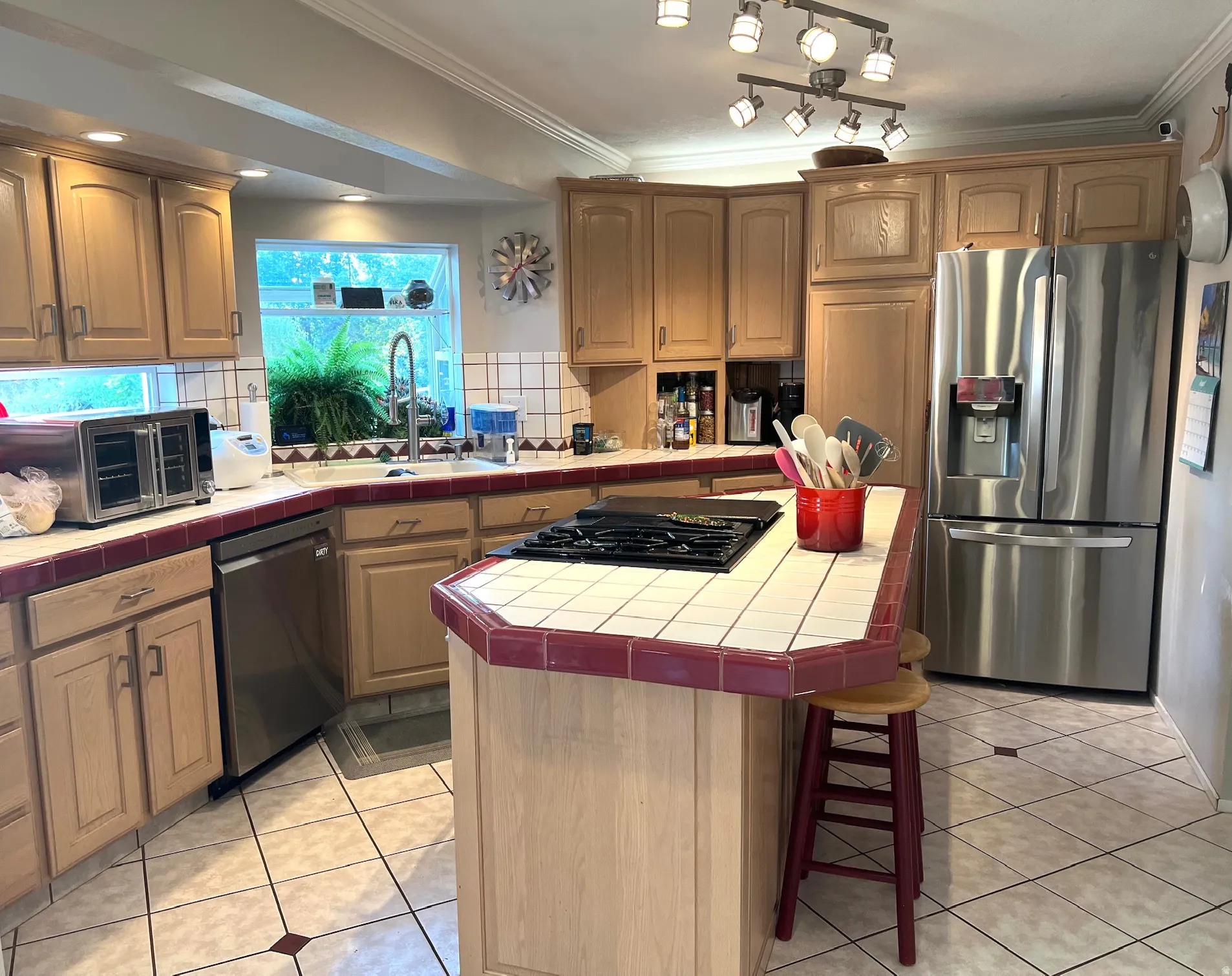 Kitchen featuring light wood cabinetry, a tile countertop with a burgundy trim, a stainless steel refrigerator, a built-in cooktop on a central island, and recessed lighting
