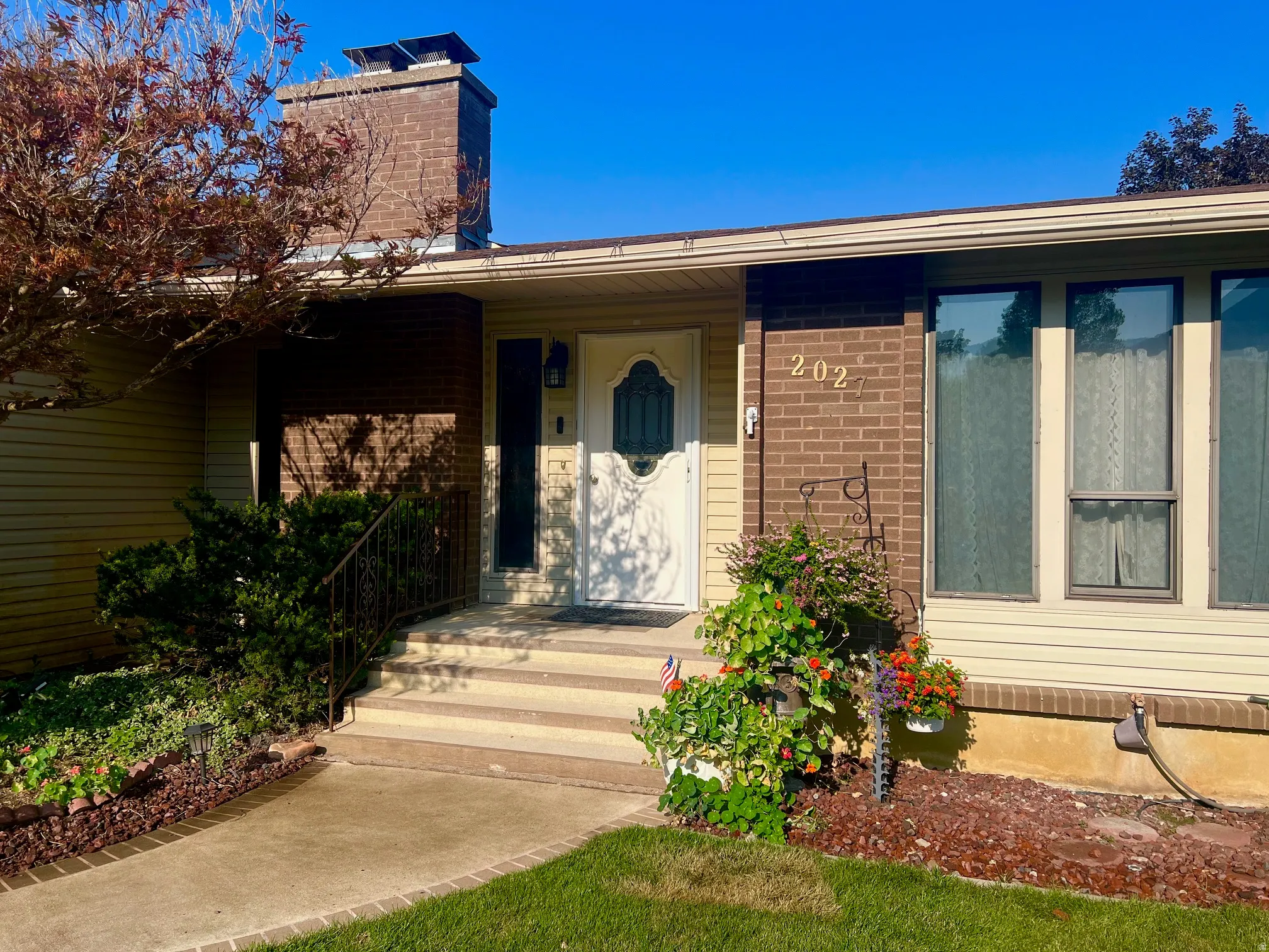 Welcoming front entry featuring a brick exterior with chimney, covered porch with concrete walkway, large picture windows, and manicured landscaping with mature plantings and mulched beds
