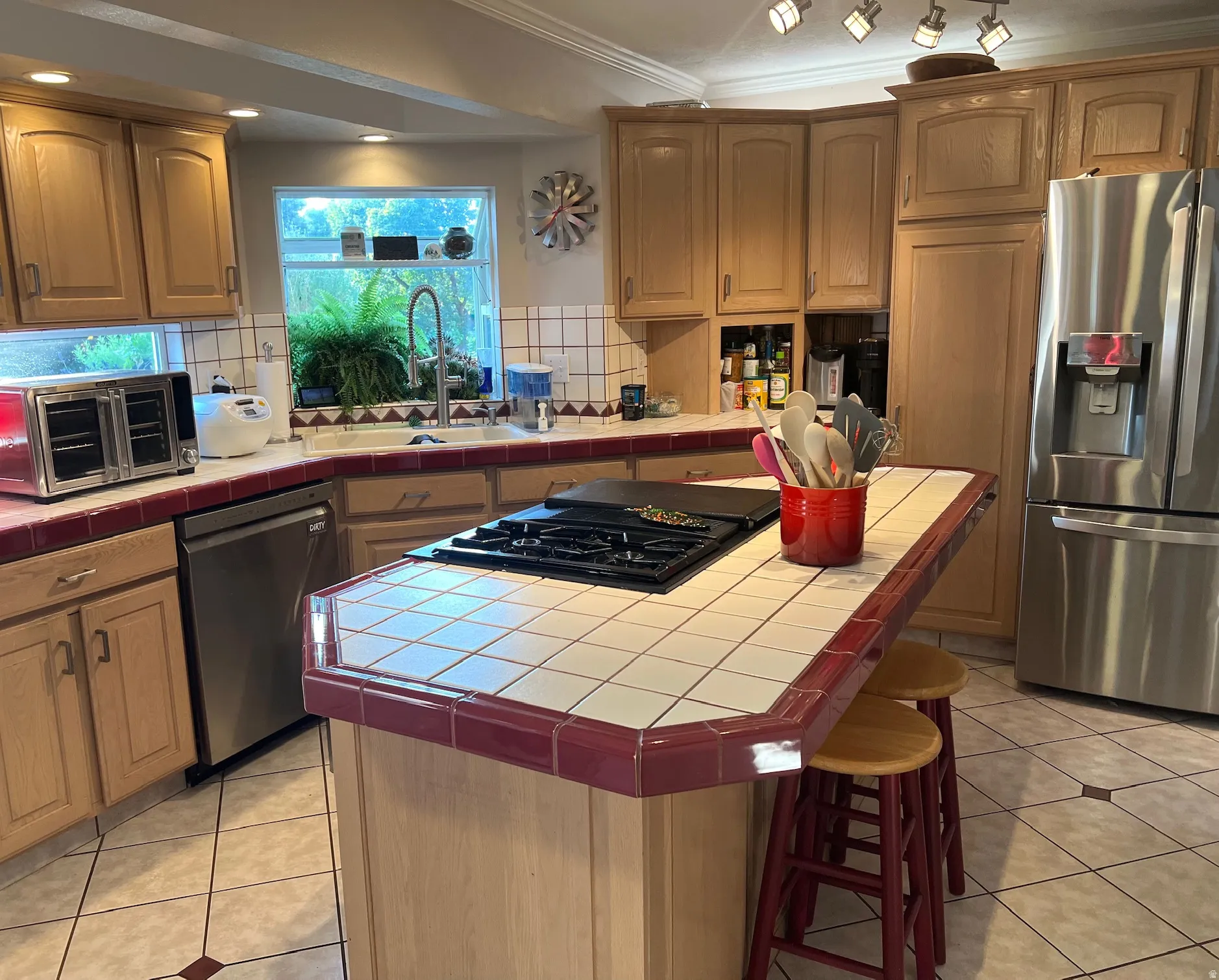 Kitchen featuring light wood cabinetry, a corner sink with a window, a stainless steel dishwasher, a stainless steel refrigerator, and a tile-topped island with an integrated cooktop