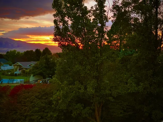 Mature tree-lined grounds with expansive lawn, sunset mountain backdrop, and detached outbuildings including pool house structure