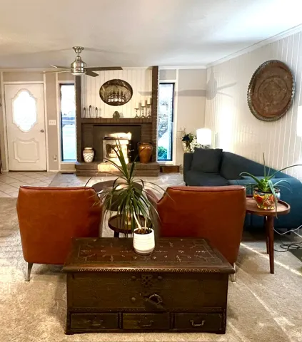 Elegant living room showcasing warm wood-finish flooring, a decorative fireplace with dark surround and mantelpiece, ceiling fan, and abundant natural light from multiple windows and glass door entry