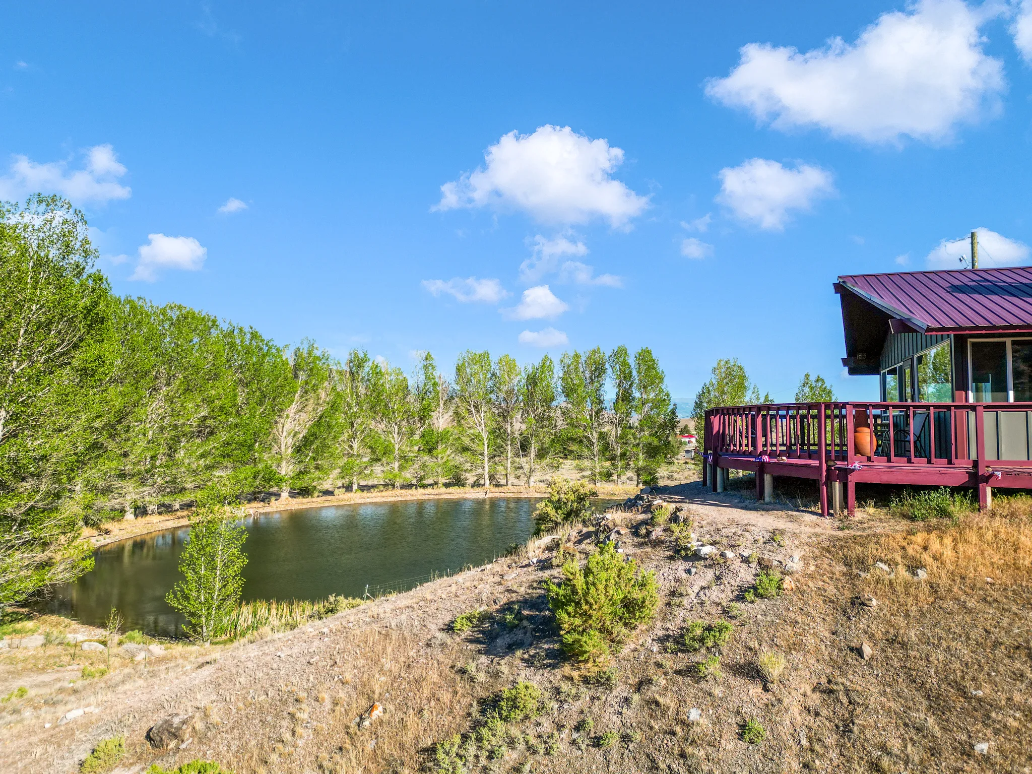 Exterior deck with wood railing, large pond, mature trees, and a metal roof