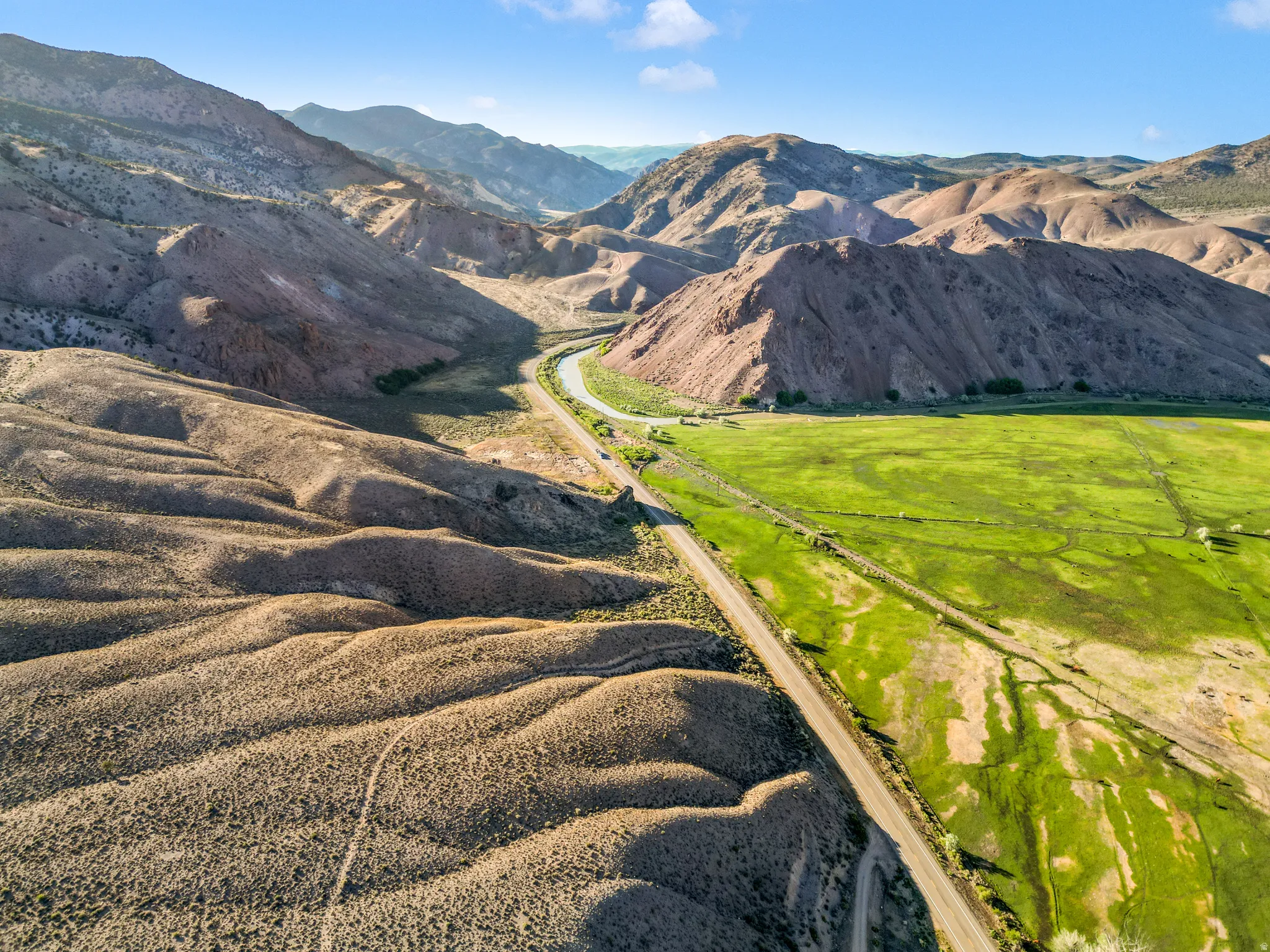 Expansive natural landscape with rolling hills, a winding road, and vibrant green fields, surrounded by rugged mountain terrain