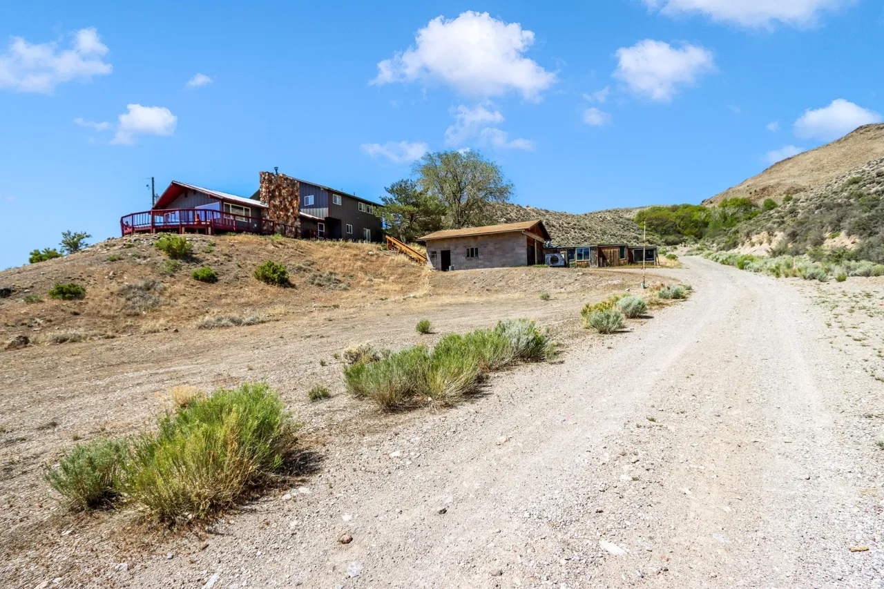 Hillside residence featuring a full-width deck with red railings, a prominent stone chimney, and a gravel driveway leading to additional structures