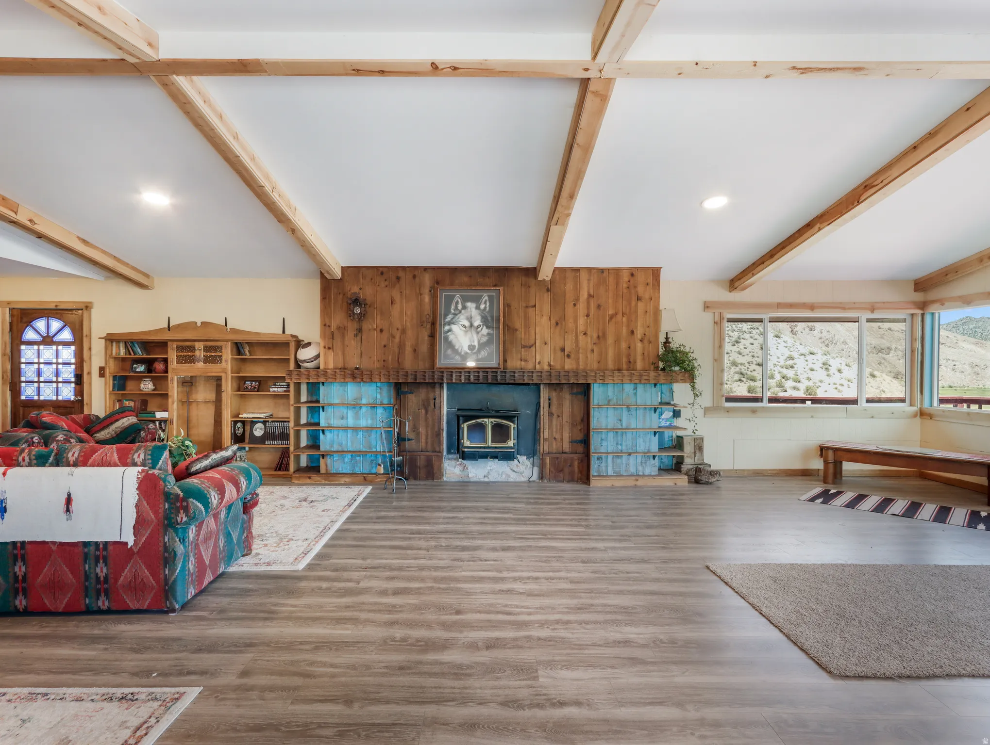 Spacious living area featuring wood-finish flooring, exposed ceiling beams, a wood-paneled accent wall, a built-in fireplace, and large windows