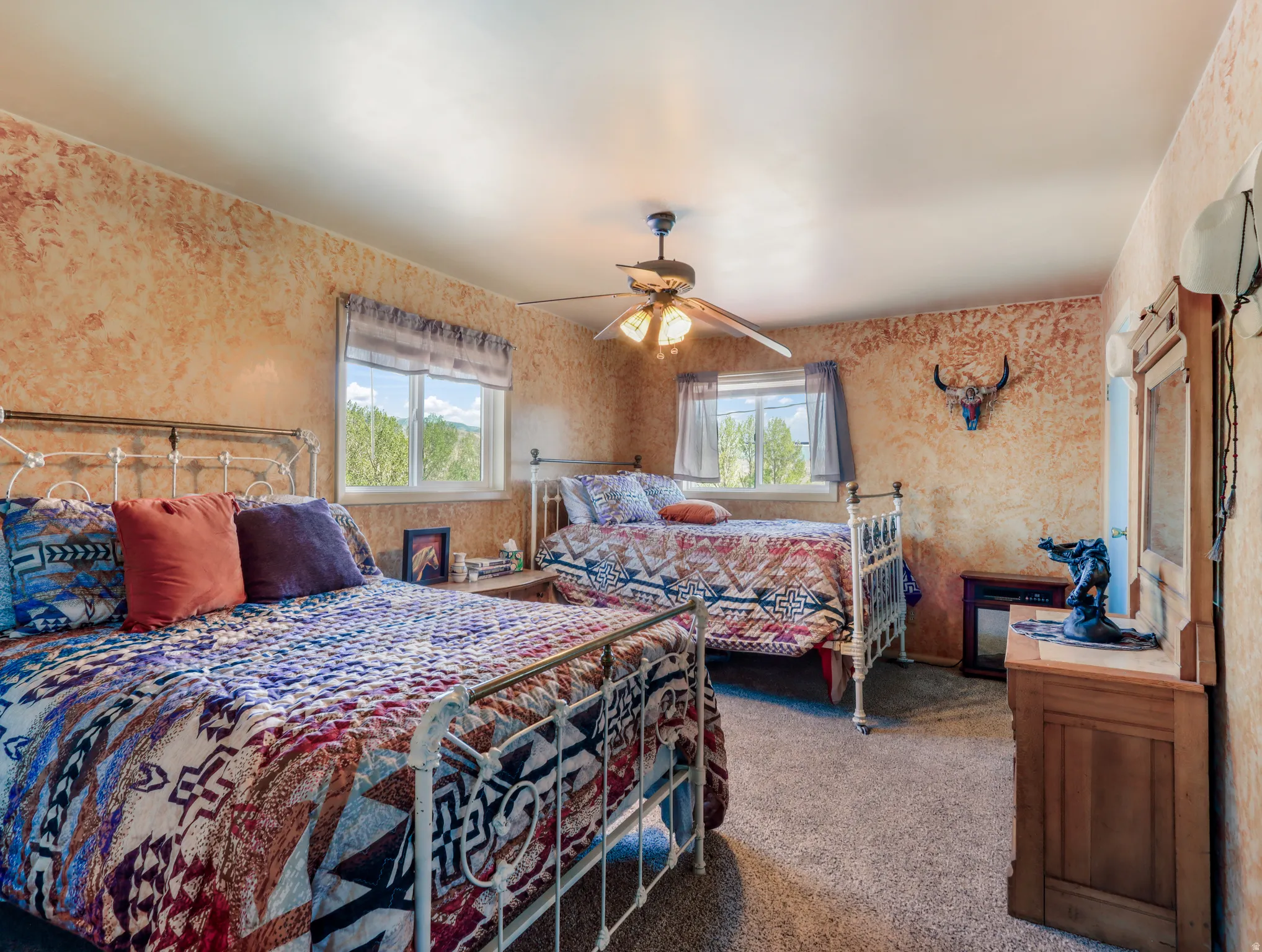 Textured wall finish, ceiling fan with light fixture, two windows with roller blinds, carpeted flooring, and a wood-finish dresser