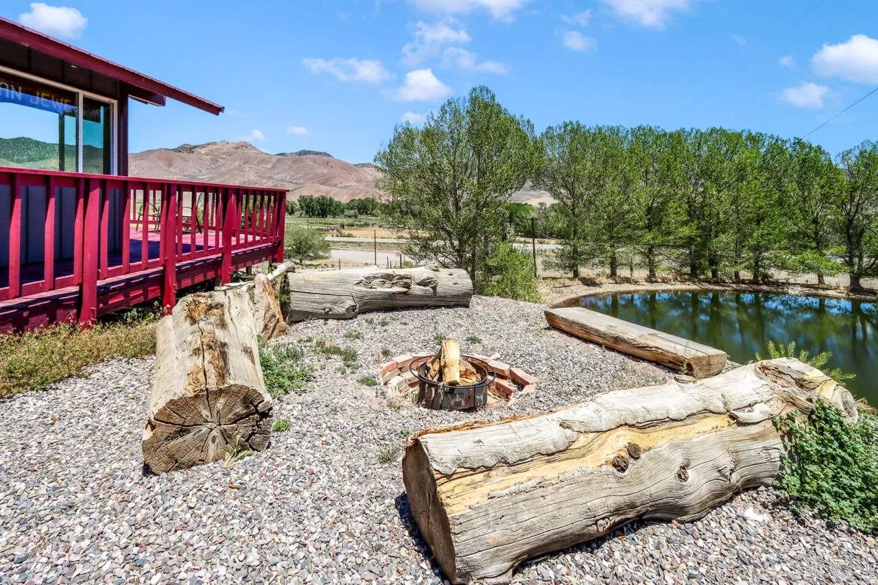 Elevated deck with red railing, gravel ground cover, fire pit, natural log seating, and a water feature surrounded by mature trees