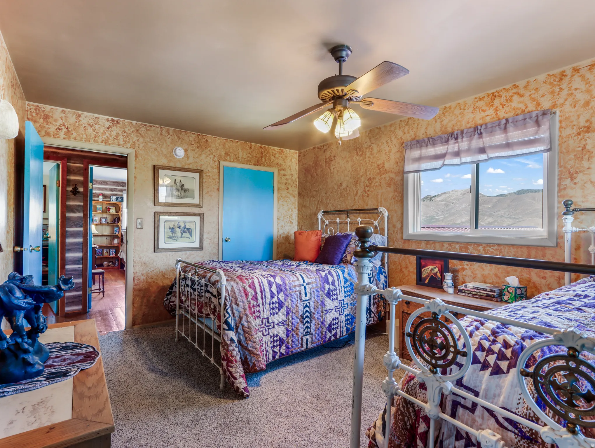 Carpeted room with a ceiling fan, a window with distant scenery, and two beds with decorative metal frames