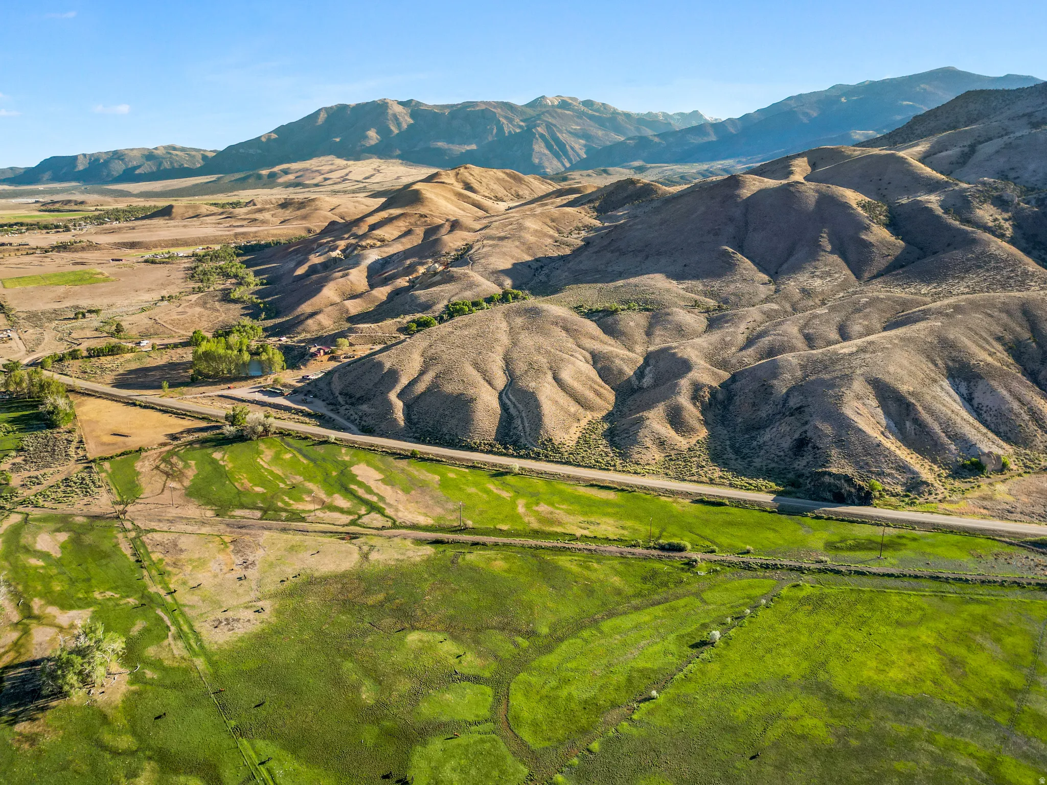 Expansive green fields with a winding road, surrounded by rolling hills and distant mountains under a clear sky
