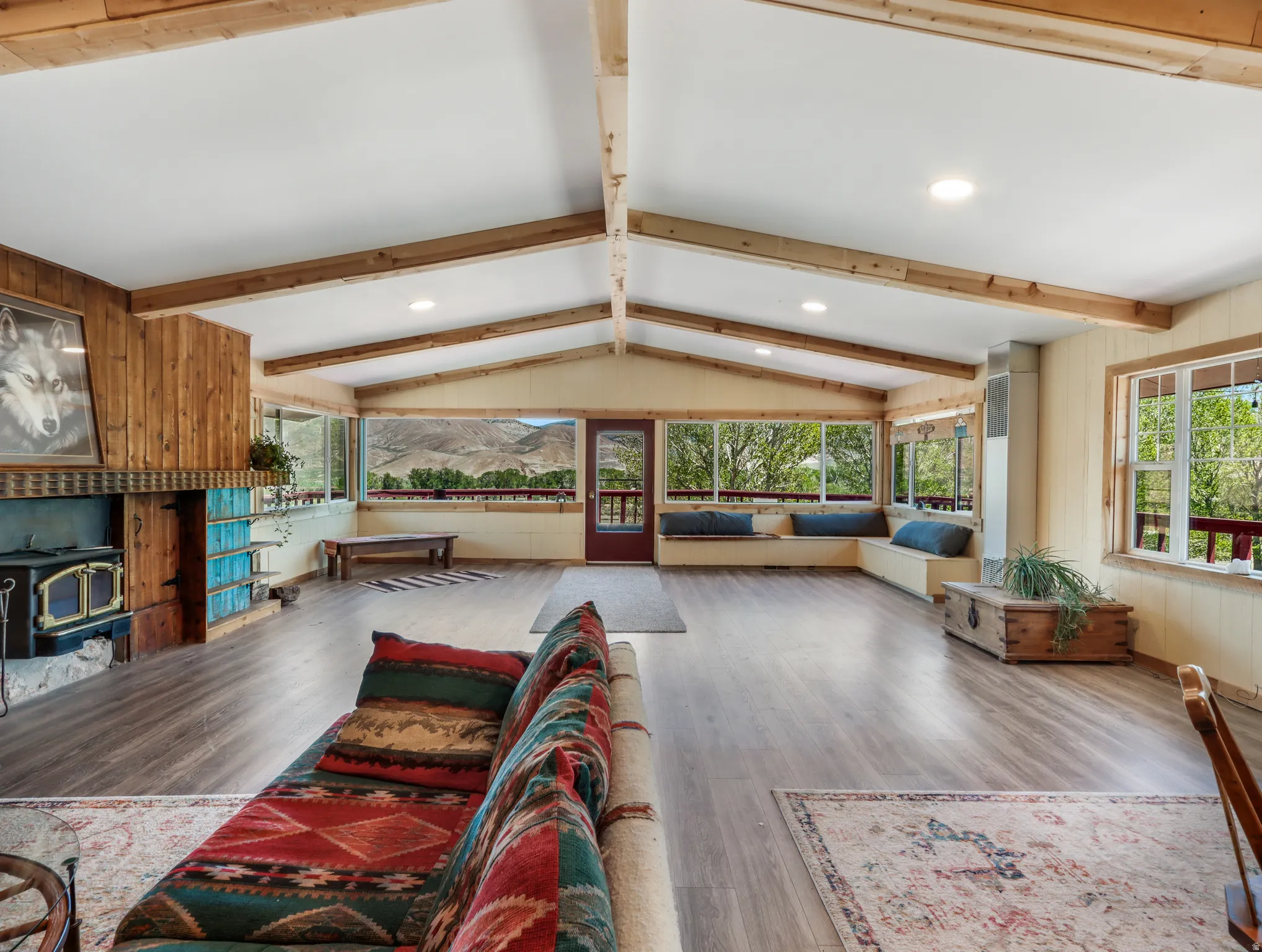Vaulted ceiling with exposed wood beams, wood-finish flooring, wood-paneled accent wall, built-in window seating, and an integrated wood-burning stove