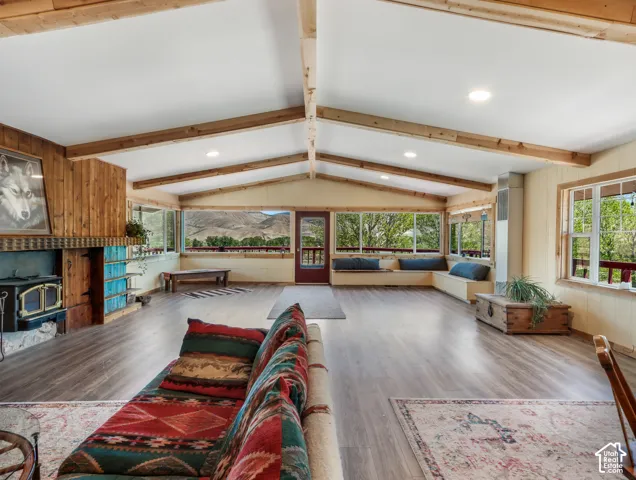 Vaulted ceiling with exposed wood beams, wood-finish flooring, wood-paneled accent wall, built-in window seating, and an integrated wood-burning stove