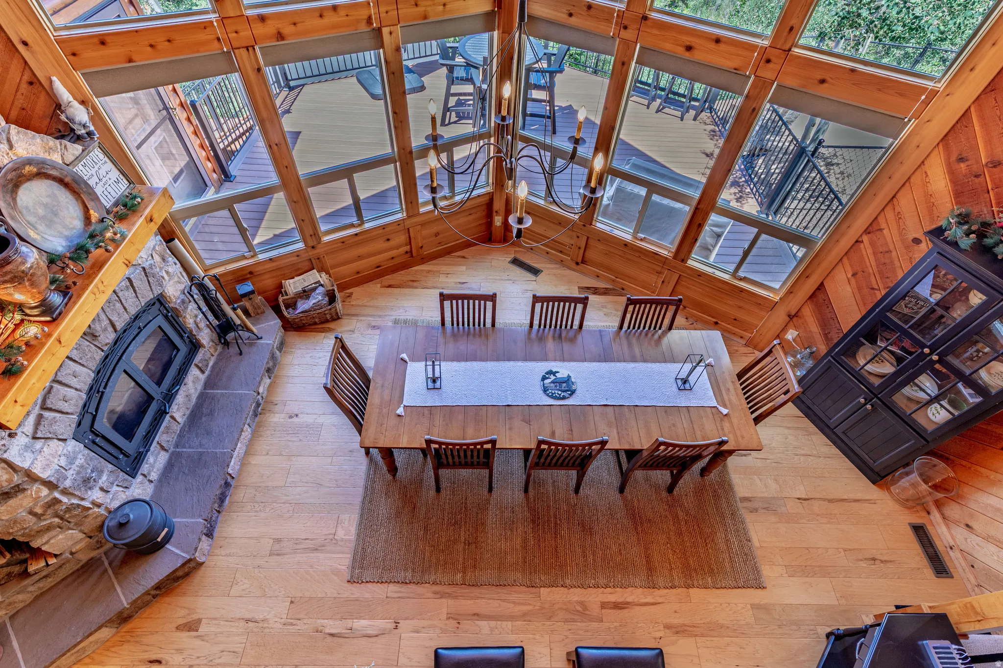 Vaulted ceiling with exposed wood beams, expansive windows, wood-finish flooring, stone fireplace, and a chandelier