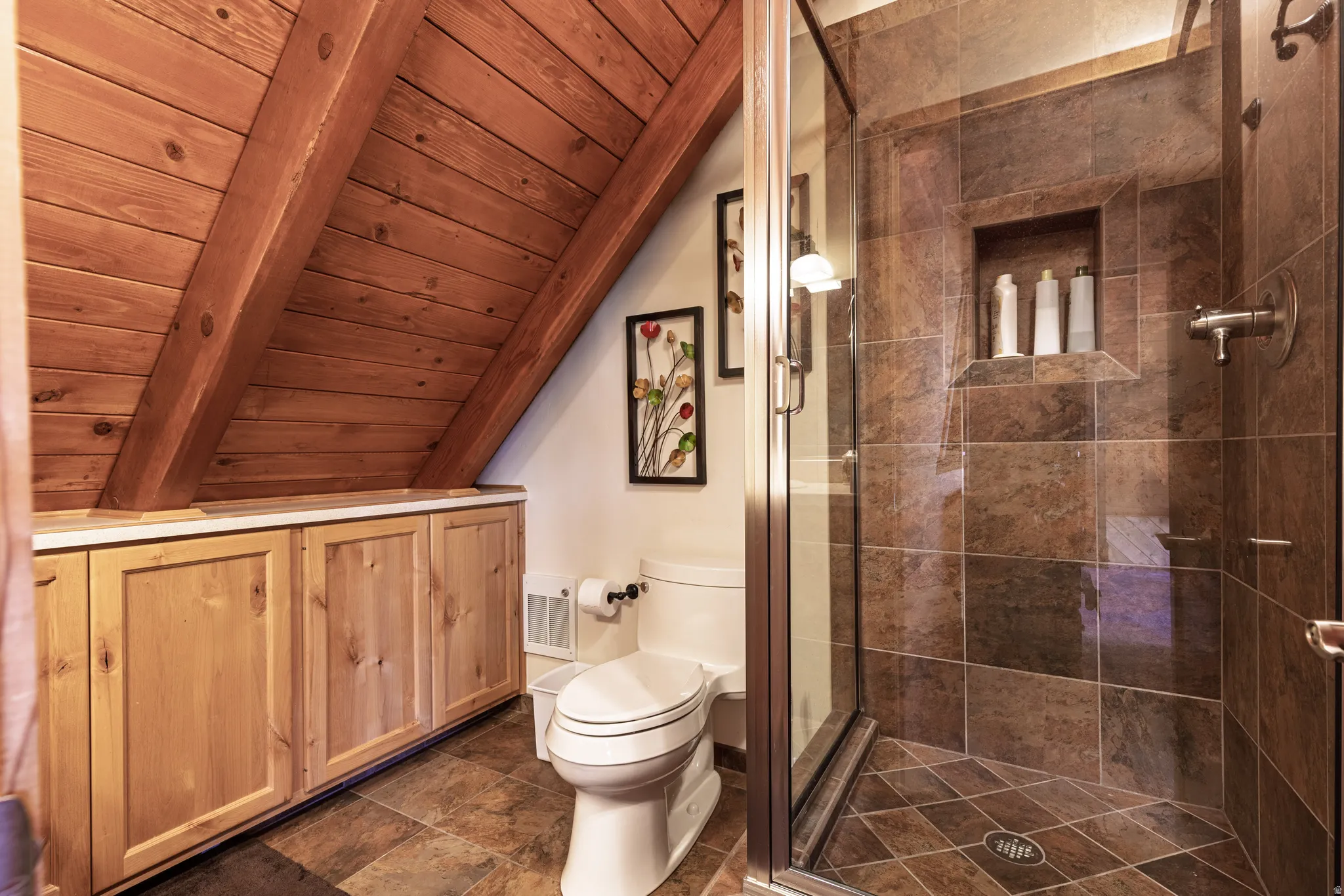 Sloped wood-paneled ceiling, built-in natural wood cabinetry with white countertop, full glass-enclosed shower with tile surround, built-in shower niche, and coordinating tile flooring