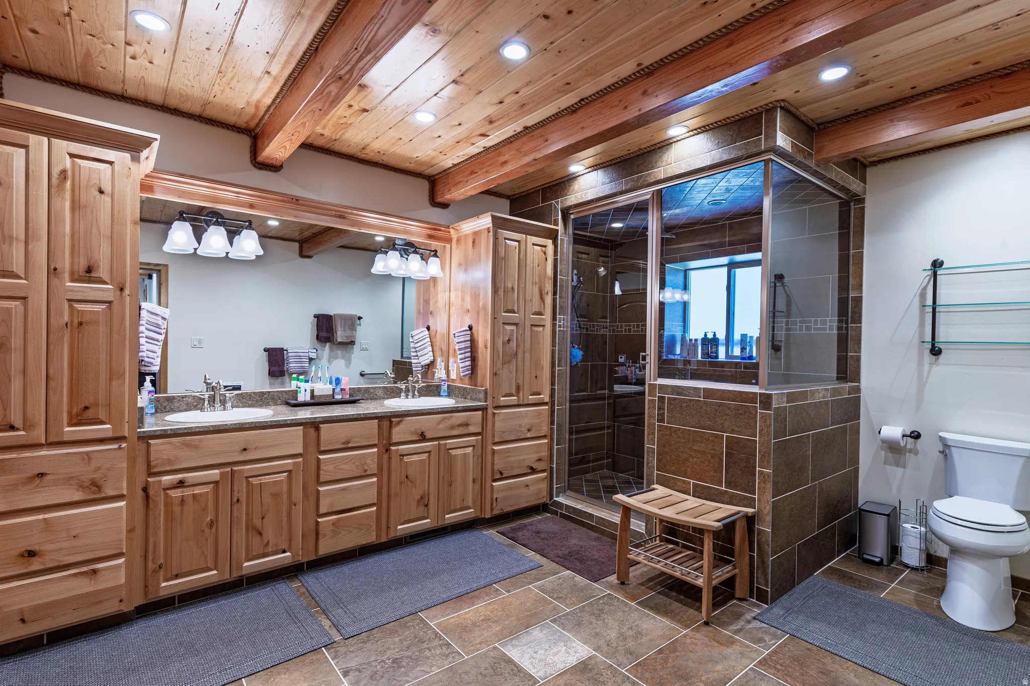 Wood-paneled ceiling with exposed beams, dual vanity with undermount sinks, and a glass-enclosed shower with tiled surround