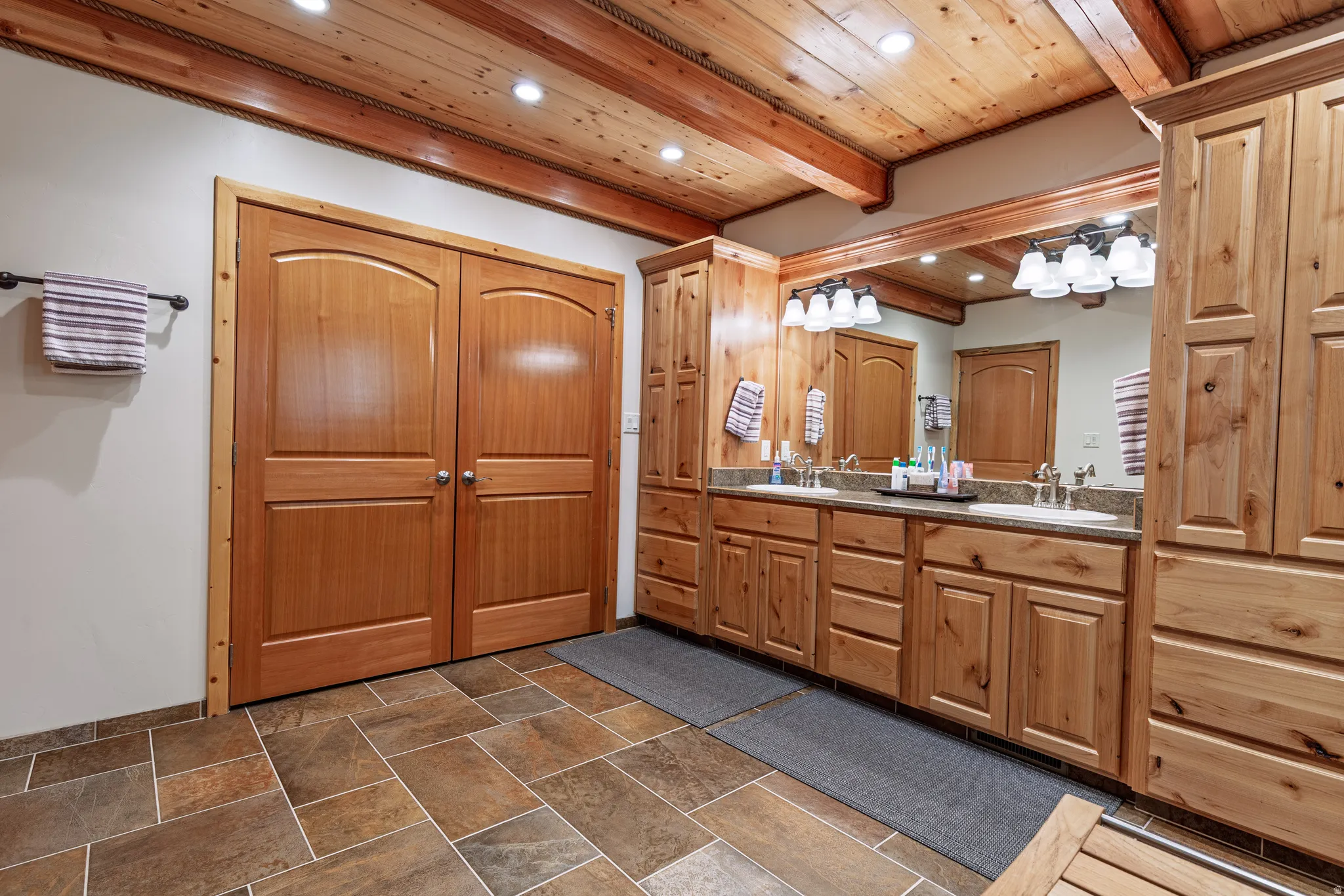 Wood-finish ceiling with exposed beams, recessed lighting, dual vanity with undermount sinks, and tile flooring