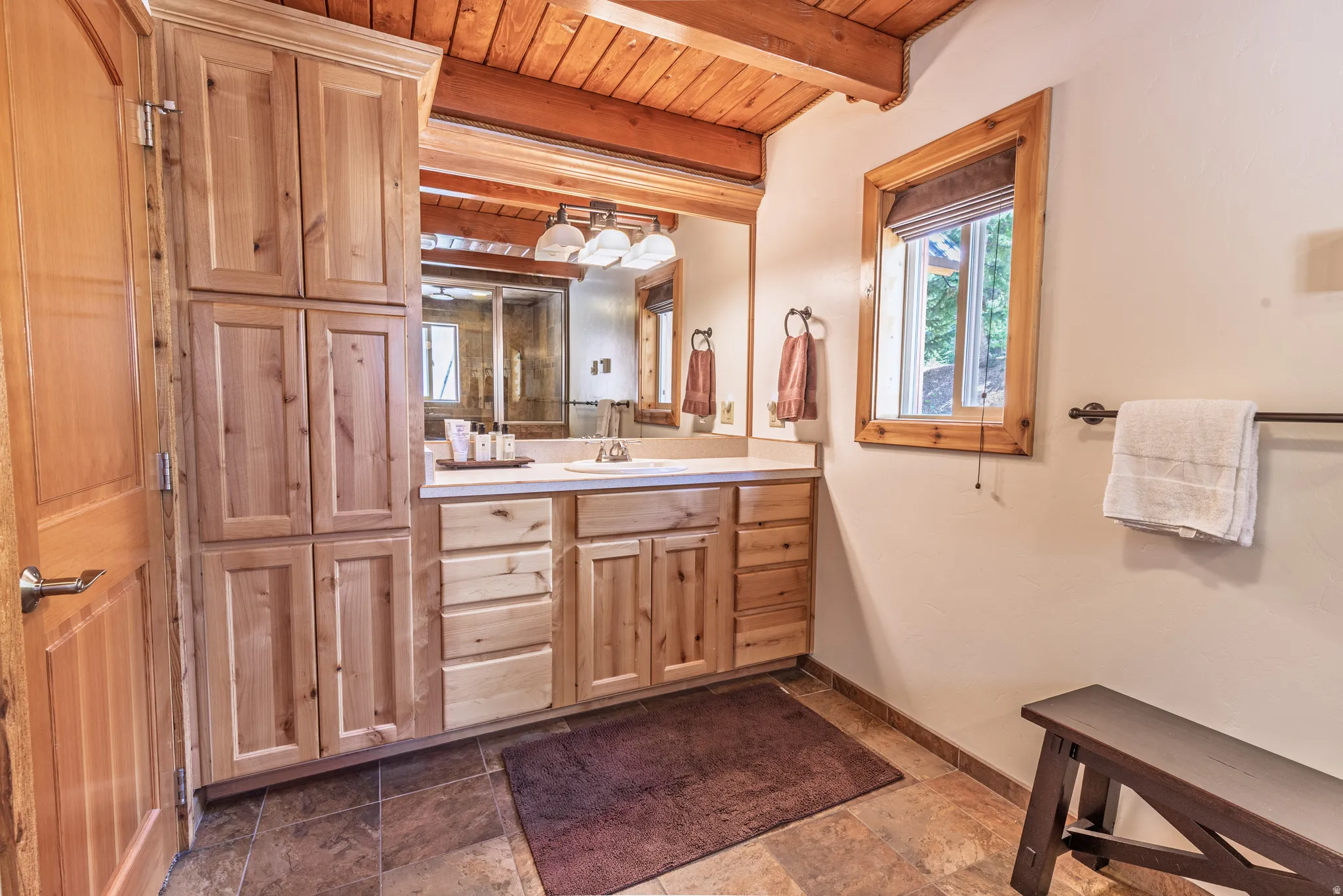 Wood-finish cabinetry, stone tile flooring, vanity with a light-colored countertop, wood-paneled ceiling, and a framed window
