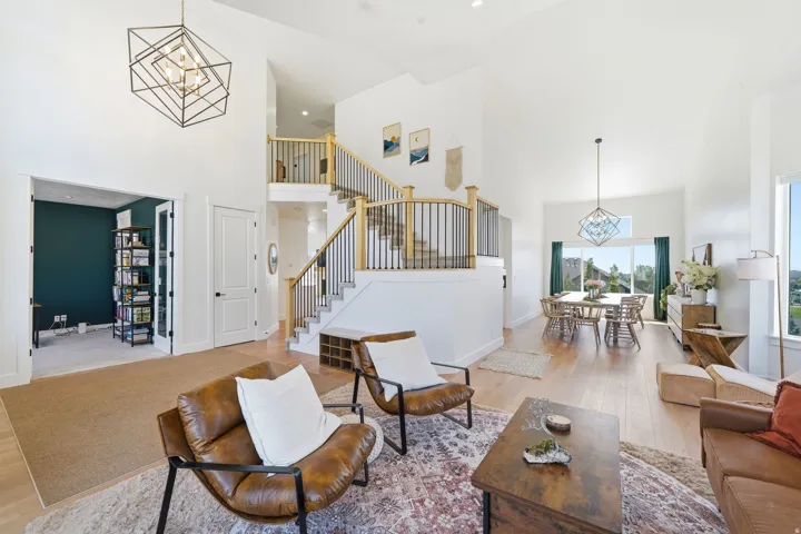 Open-concept living space anchored by light wood-finish flooring throughout, featuring a geometric pendant light fixture, a lofted mezzanine with metal railings, and a dark green accent wall with built-in shelving