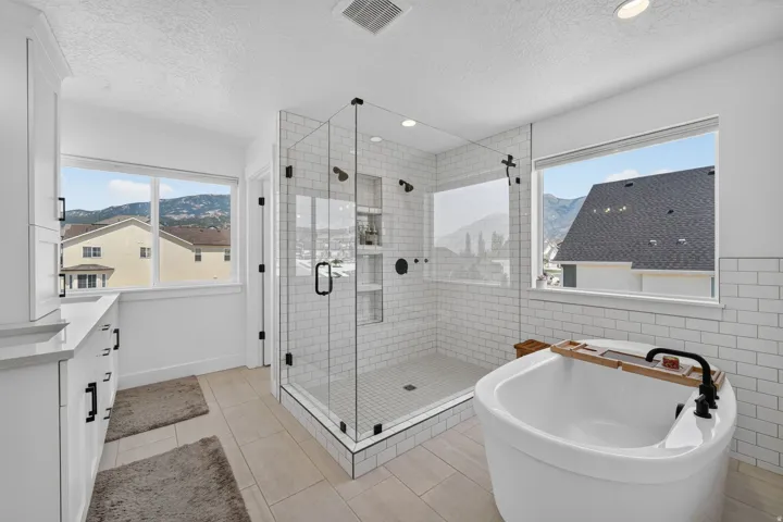 Freestanding soaking tub, glass-enclosed shower with subway tile, dual vanity with white cabinetry, and large windows