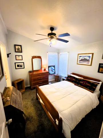 Primary bedroom with vaulted ceiling, ceiling fan with integrated lighting, textured wall finishes, carpeted flooring, and two windows with blinds.