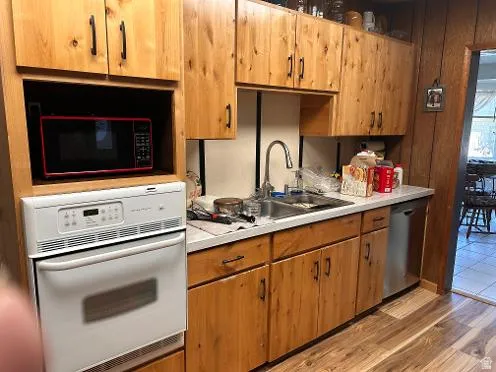 Wood-finish flooring, wood cabinetry with black hardware, white countertops, built-in white oven, and stainless steel dishwasher