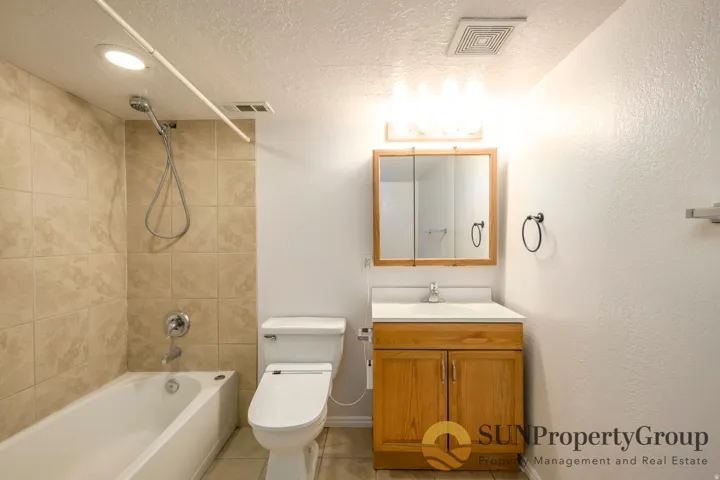 Bathtub with tiled surround, wood-finish vanity with countertop sink, and wall-mounted medicine cabinet with integrated lighting