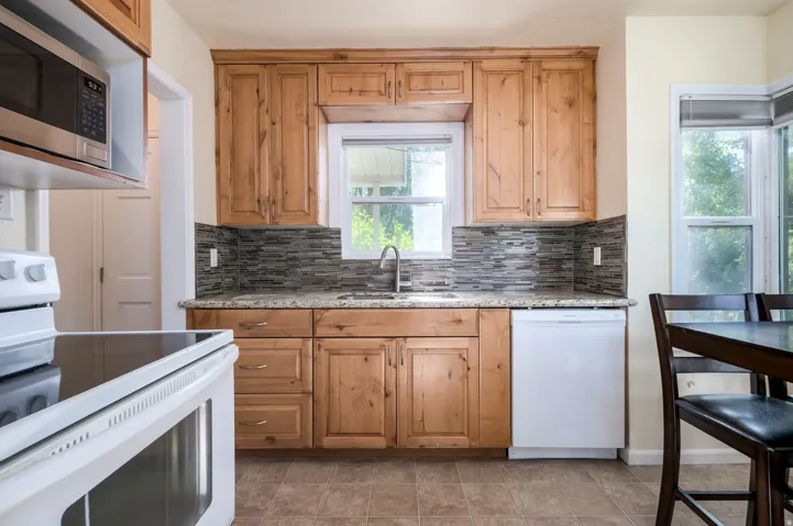 Wood-finish cabinetry, granite-finish countertops, mosaic tile backsplash, stainless steel microwave, and white appliances