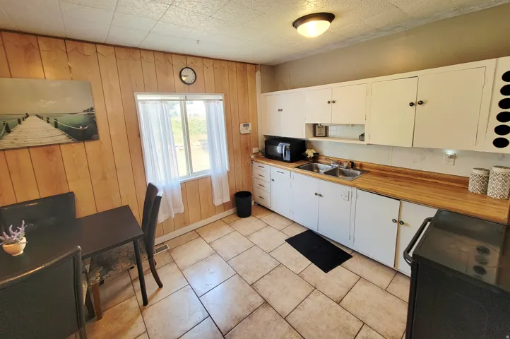 Wood-paneled wall, white upper and lower cabinetry, wood-finish countertops, stainless steel double-basin sink, and tiled flooring