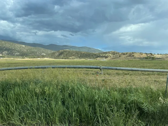 Expansive natural landscape with mountain backdrop and open fields, featuring a prominent irrigation pipe across the foreground