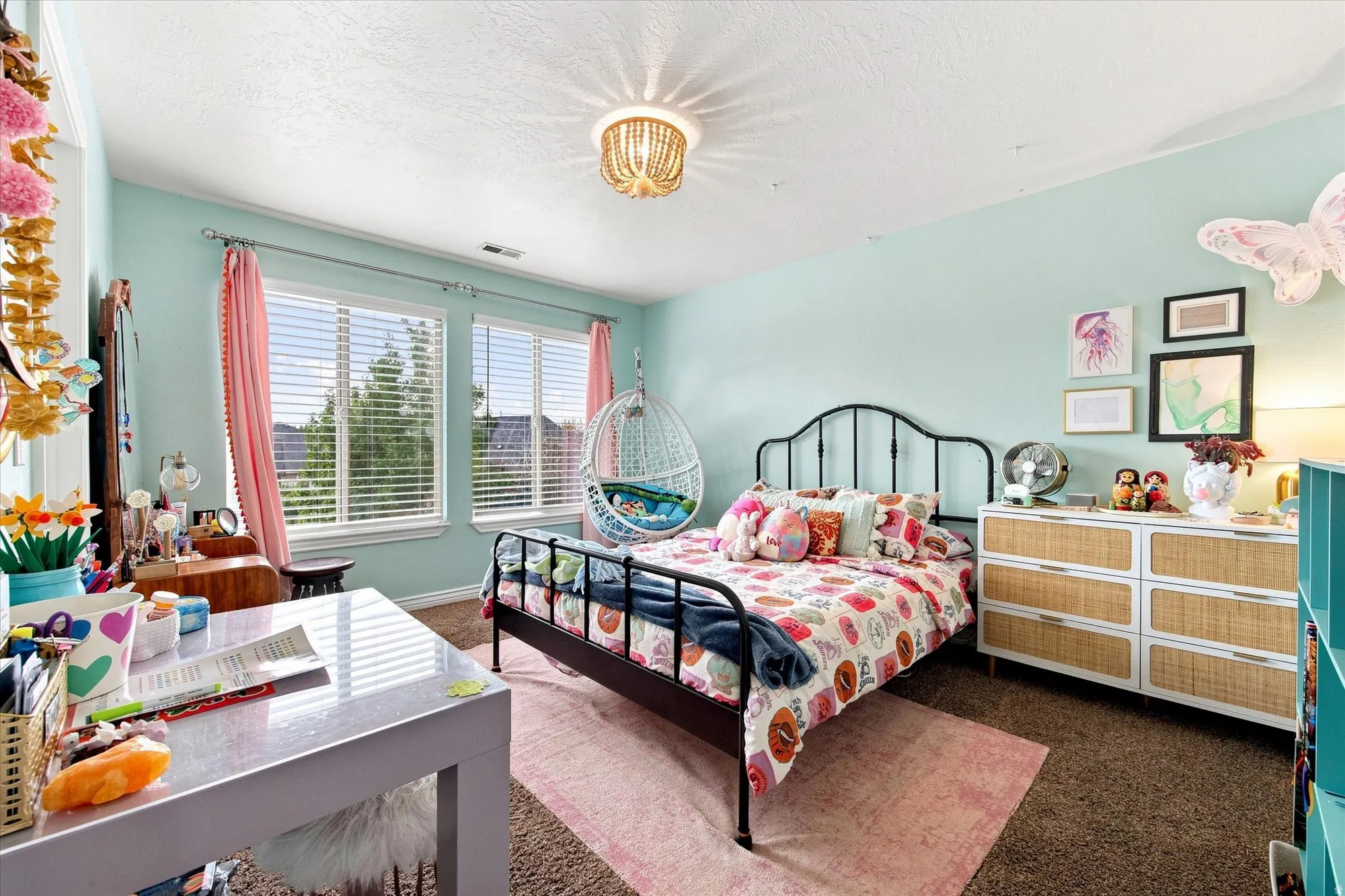 Dual windows with blinds, wall-mounted decorative light fixture, wrought iron bed frame, six-drawer dresser with cane accents, and soft wall tones