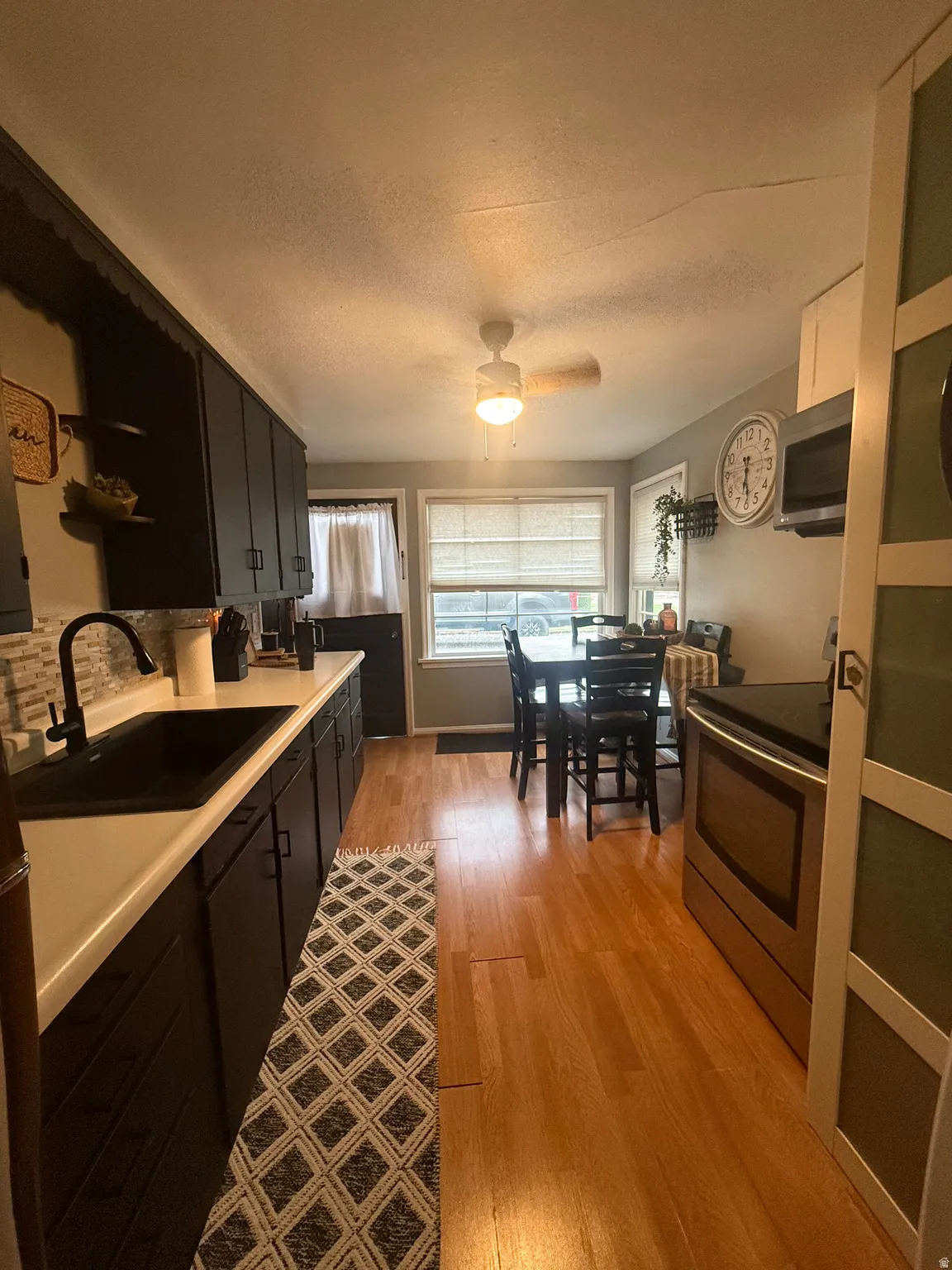 Wood-finish flooring, dark cabinetry, white countertops, stainless steel appliance, and a ceiling fan with integrated lighting