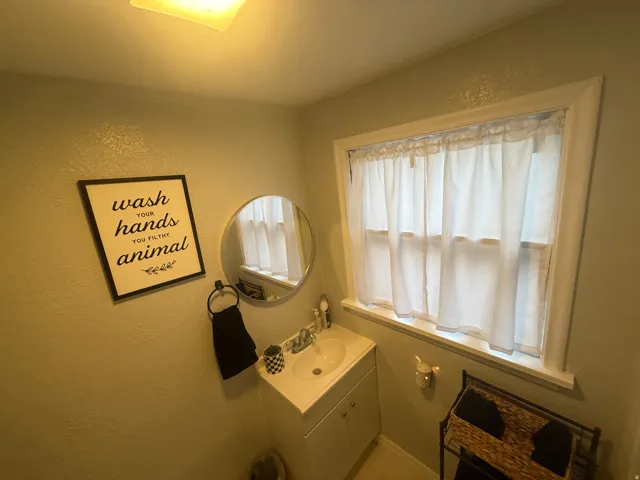 Single window with white trim, wall-mounted circular mirror, white vanity with integrated sink, and overhead lighting fixture
