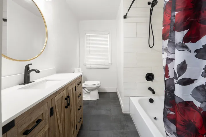 Wood-finish vanity with a white countertop and undermount sink, matte black faucet, round wall-mounted mirror, and a white tiled shower/tub combination