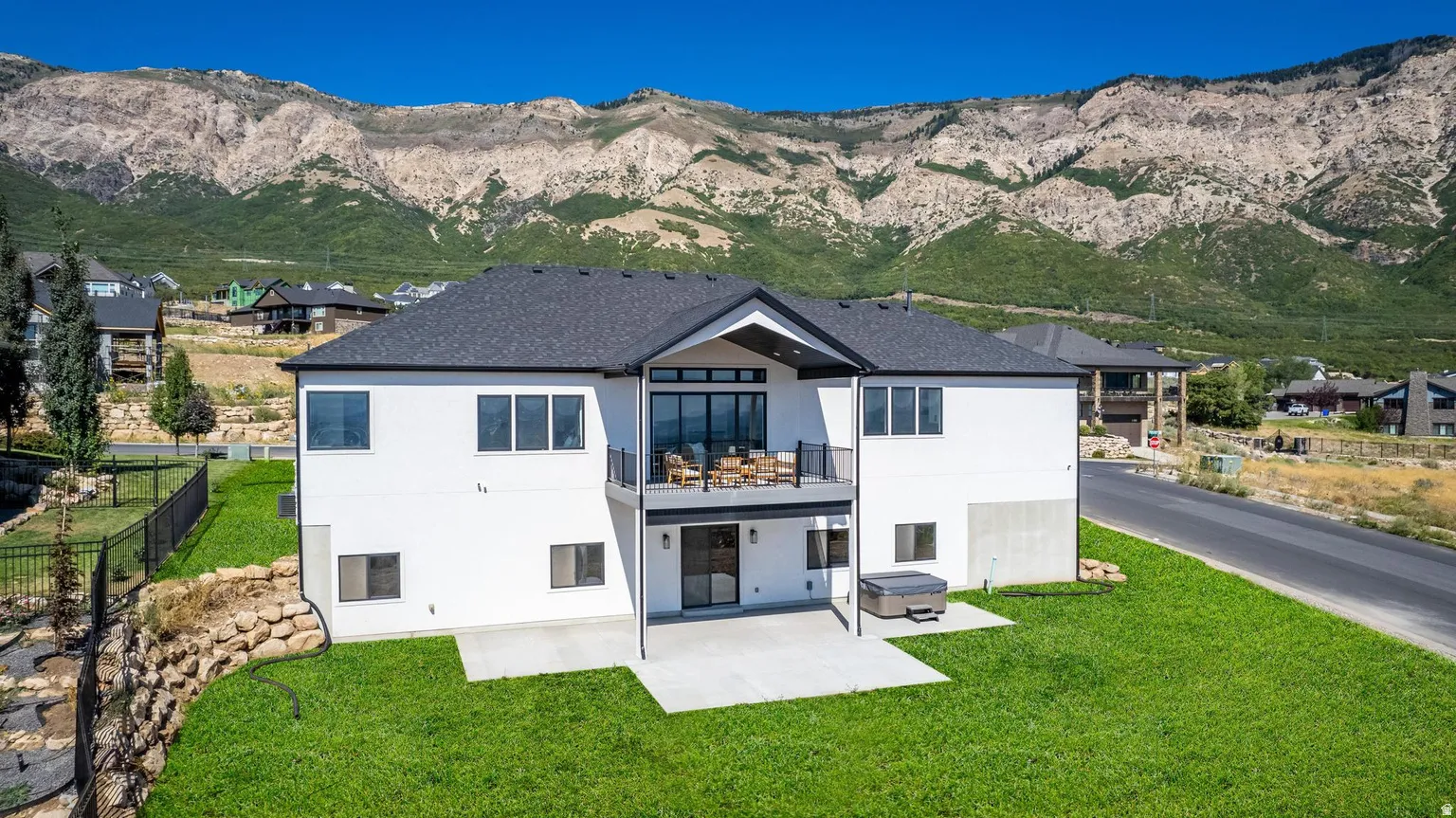 White exterior residence featuring a dark shingle roof, upper-level balcony, lower-level patio, and a backyard hot tub