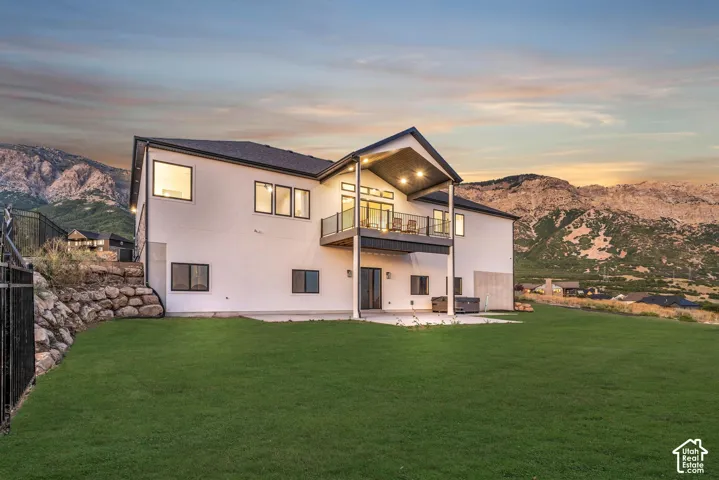 Expansive green lawn, multi-level patio, upper-level balcony with glass railing, mountain backdrop, and contemporary exterior finish