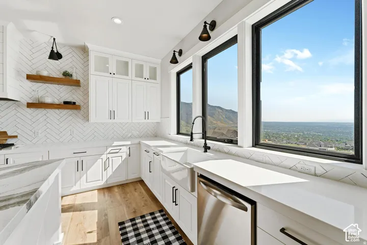 White shaker-style cabinetry with matte black hardware, herringbone tile backsplash, integrated farmhouse sink, stainless steel dishwasher, and wood-finish flooring