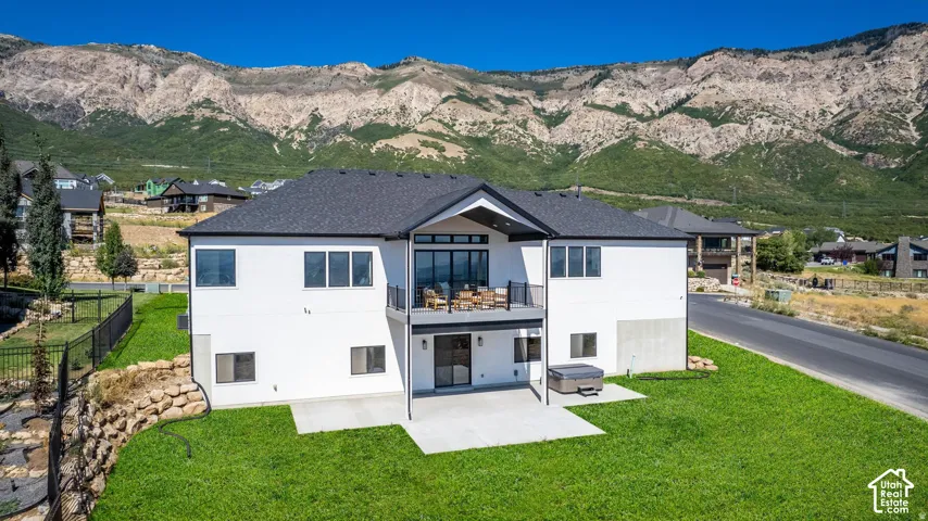 White exterior residence featuring a dark shingle roof, upper-level balcony, lower-level patio, and a backyard hot tub