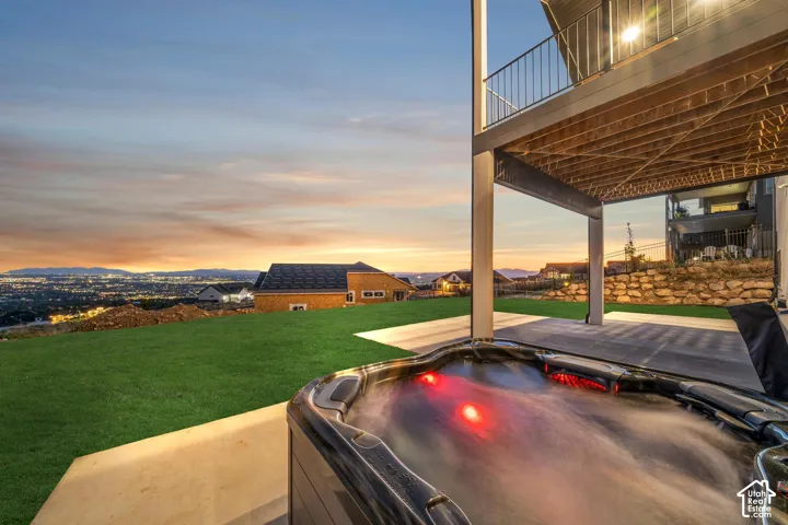 Outdoor spa with integrated lighting, expansive concrete patio, upper-level deck with metal railing, and a sloped lawn