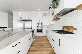 White cabinetry with black hardware, wood-finish flooring, herringbone tile backsplash, built-in stainless steel appliances, and a large central island with a stone countertop