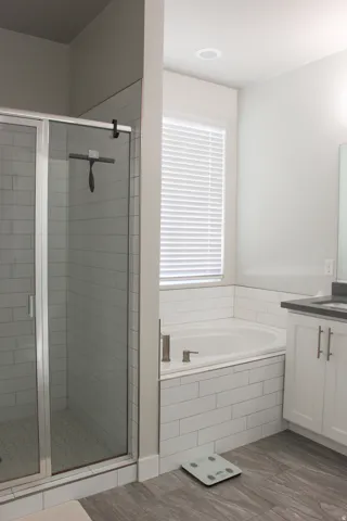 Walk-in shower with clear glass enclosure and subway tile surround, built-in soaking tub with subway tile surround, white vanity with stone countertop, and wood-finish flooring
